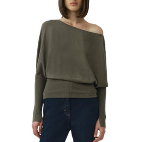 A person wears the Simkhai Lavina Draped Off Shoulder Sweater in olive-green with an asymmetric neckline, paired with dark blue jeans, standing against a plain white background.
