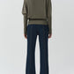 A person stands facing away, wearing the Simkhai Lavina Draped Off Shoulder Sweater in olive green and dark blue flared jeans against a plain light background.