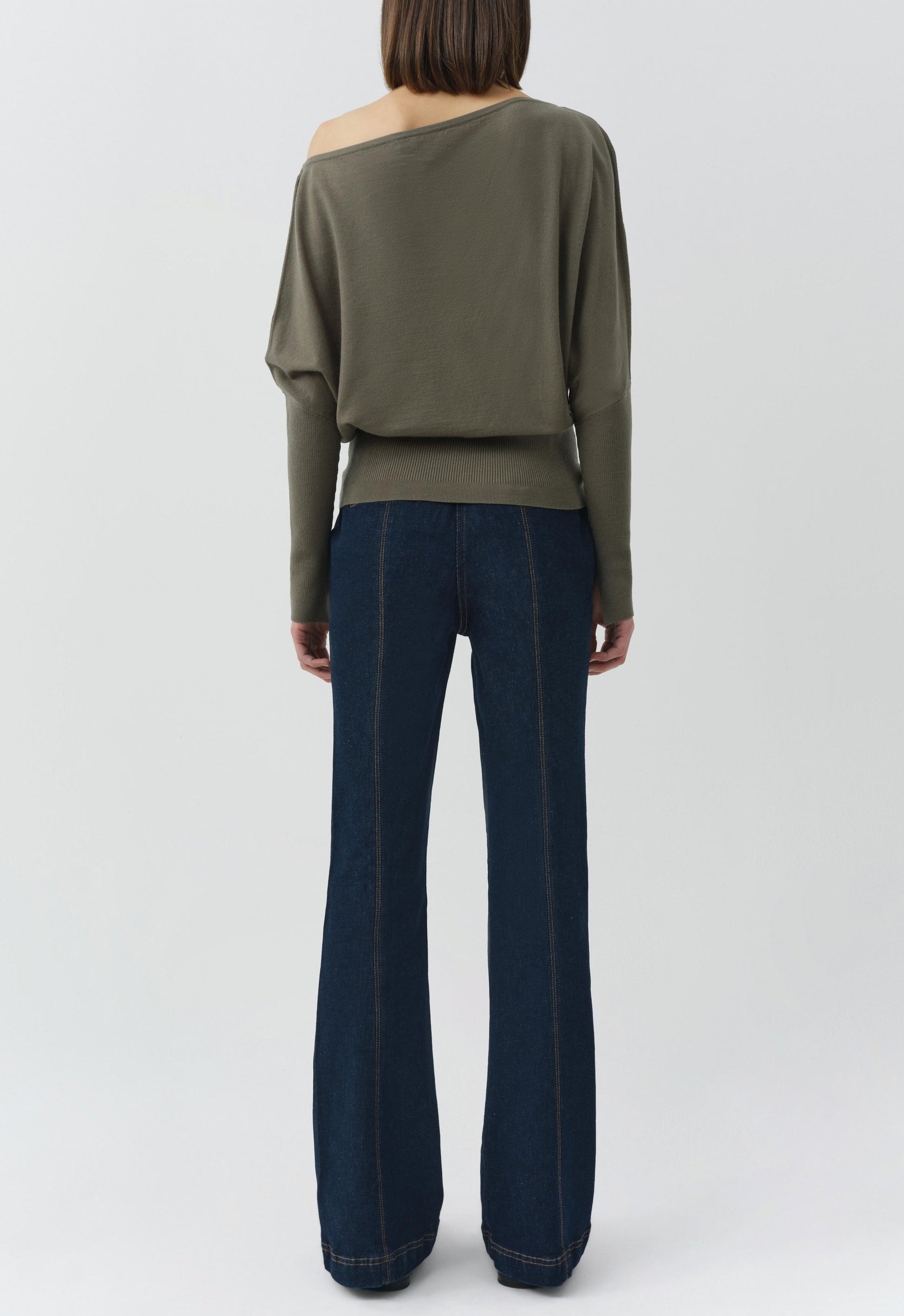 A person stands facing away, wearing the Simkhai Lavina Draped Off Shoulder Sweater in olive green and dark blue flared jeans against a plain light background.