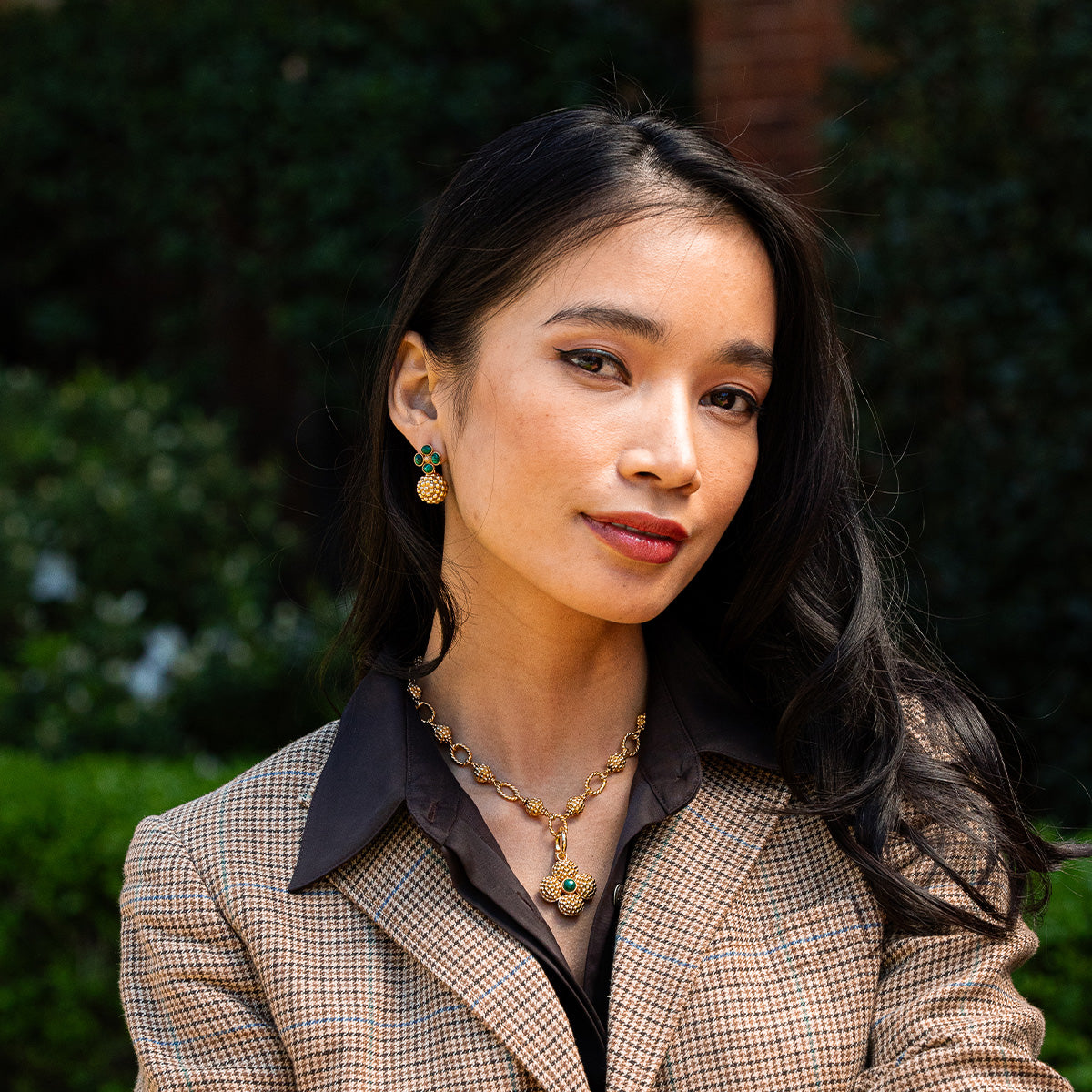 A woman with long dark hair wears a brown plaid blazer, black shirt, and gold jewelry, including Juliska Capucine De Wulf's Berry Clover Double Sided Pendant necklace, standing outdoors with greenery in the background.