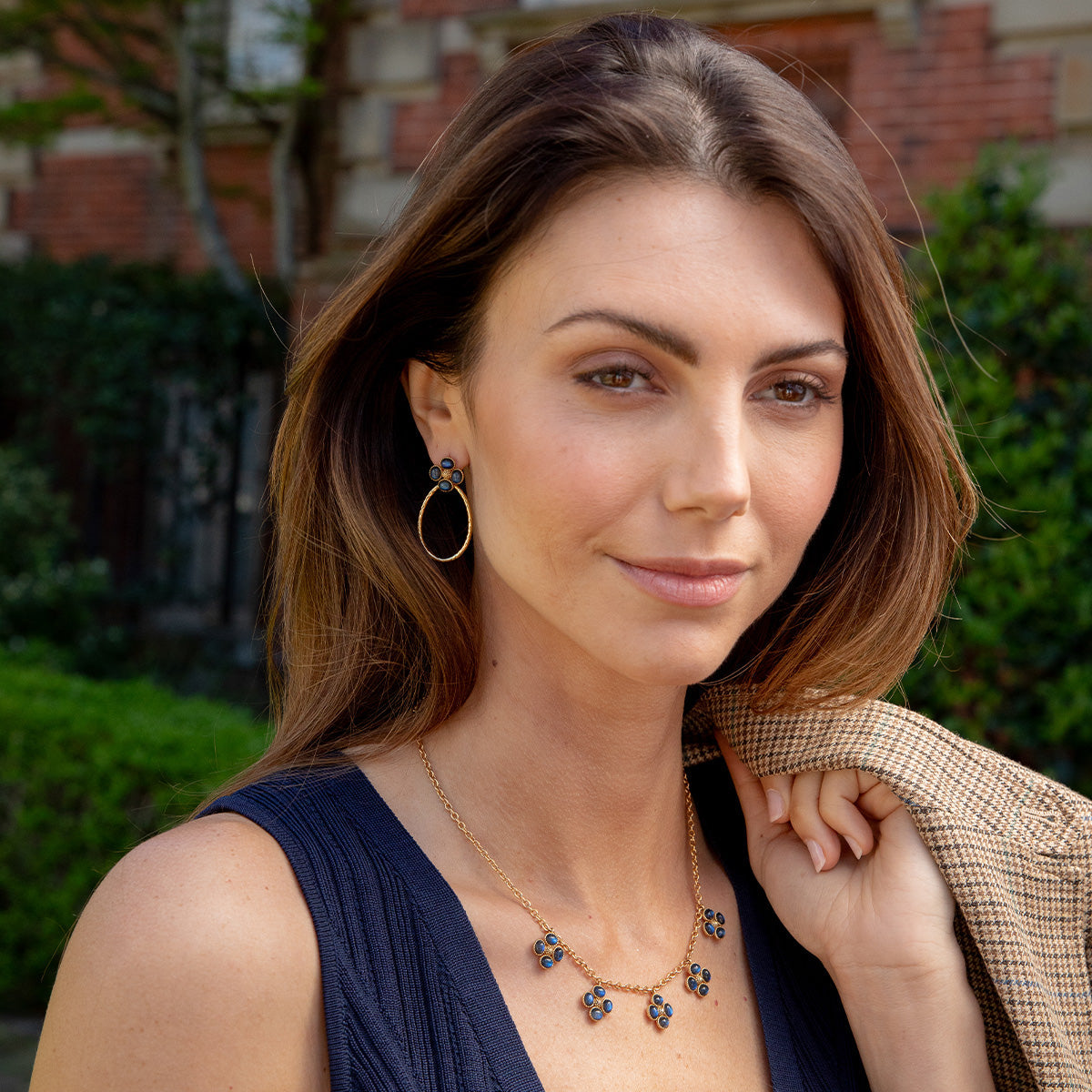 A woman with long brown hair wears a navy sleeveless top and Juliska Capucine De Wulf Berry Clover Teardrop Earrings, holding a tan blazer over her shoulder while standing outdoors.