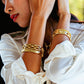 A woman in a white blouse wears the Juliska Capucine De Wulf Berry Classic Cuff and several gold bracelets on her wrist, with her arms raised near her face.