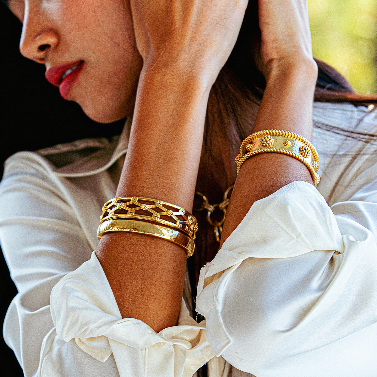 A woman in a white blouse wears the Juliska Capucine De Wulf Berry Classic Cuff and several gold bracelets on her wrist, with her arms raised near her face.