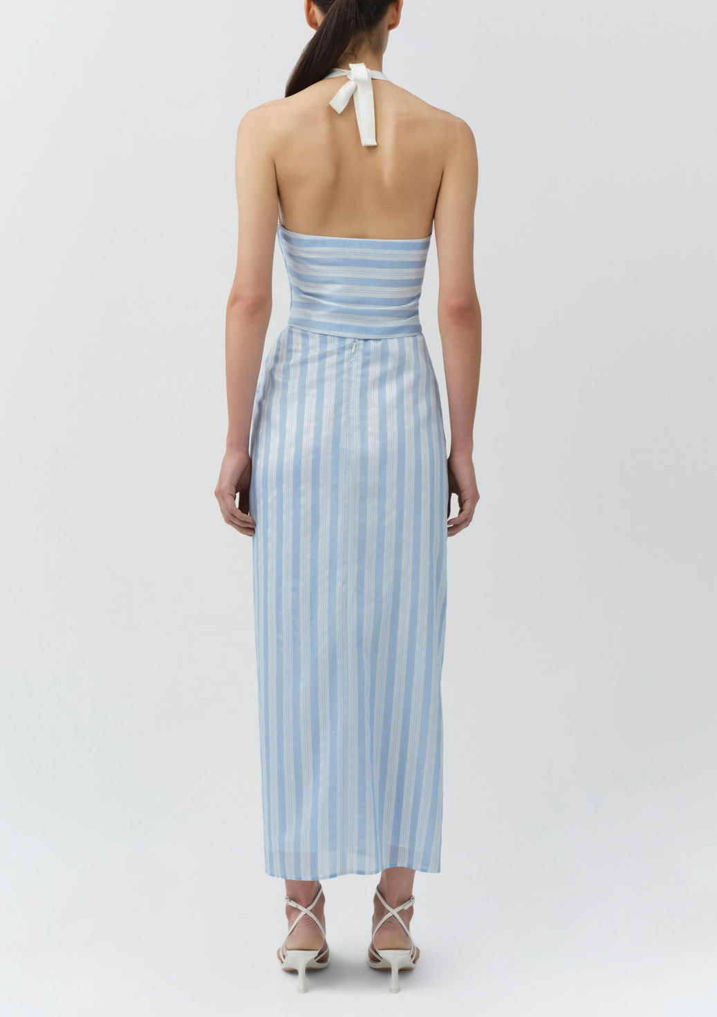 A woman stands with her back to the camera, wearing the Simkhai Caprie Halter Top in Oxford Blue and white stripes, paired with white strappy heels against a plain light background.