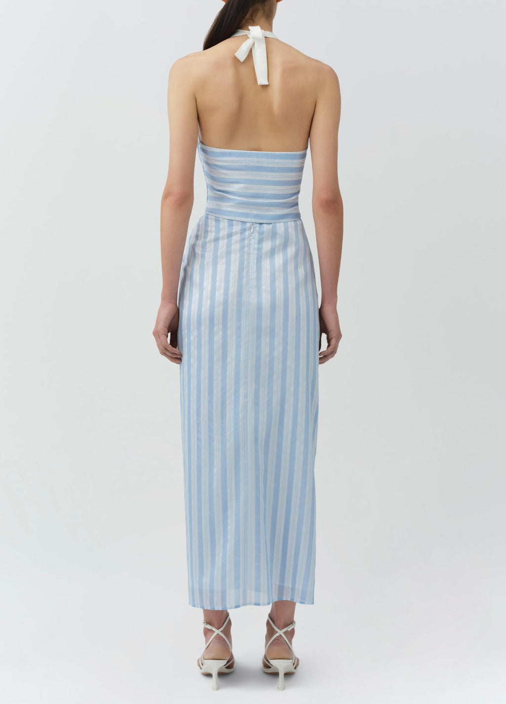 A woman faces away, wearing the Simkhai Liorra Draped Midi Skirt—a light blue and white striped wrap-effect skirt with a back tie—styled with white strappy heels.