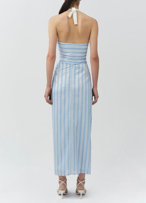 A woman faces away, wearing the Simkhai Liorra Draped Midi Skirt—a light blue and white striped wrap-effect skirt with a back tie—styled with white strappy heels.