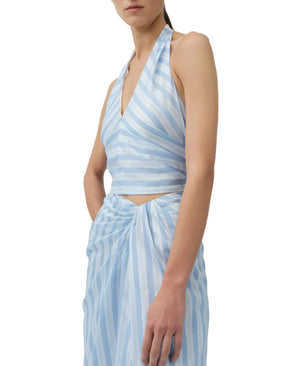 A woman models the Simkhai Caprie Halter Top in light blue and white satin stripes, paired with a matching wrap skirt. The image is cropped from shoulders to mid-thigh against a white background.