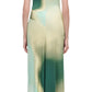 A woman stands with her back to the camera, wearing the Simkhai Acacia Jersey Midi Dress in a green and cream abstract print, featuring a sleeveless cut and exquisite drape, paired with heeled sandals.