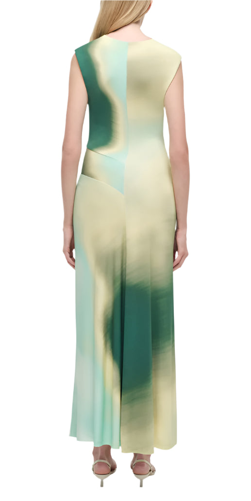 A woman stands with her back to the camera, wearing the Simkhai Acacia Jersey Midi Dress in a green and cream abstract print, featuring a sleeveless cut and exquisite drape, paired with heeled sandals.