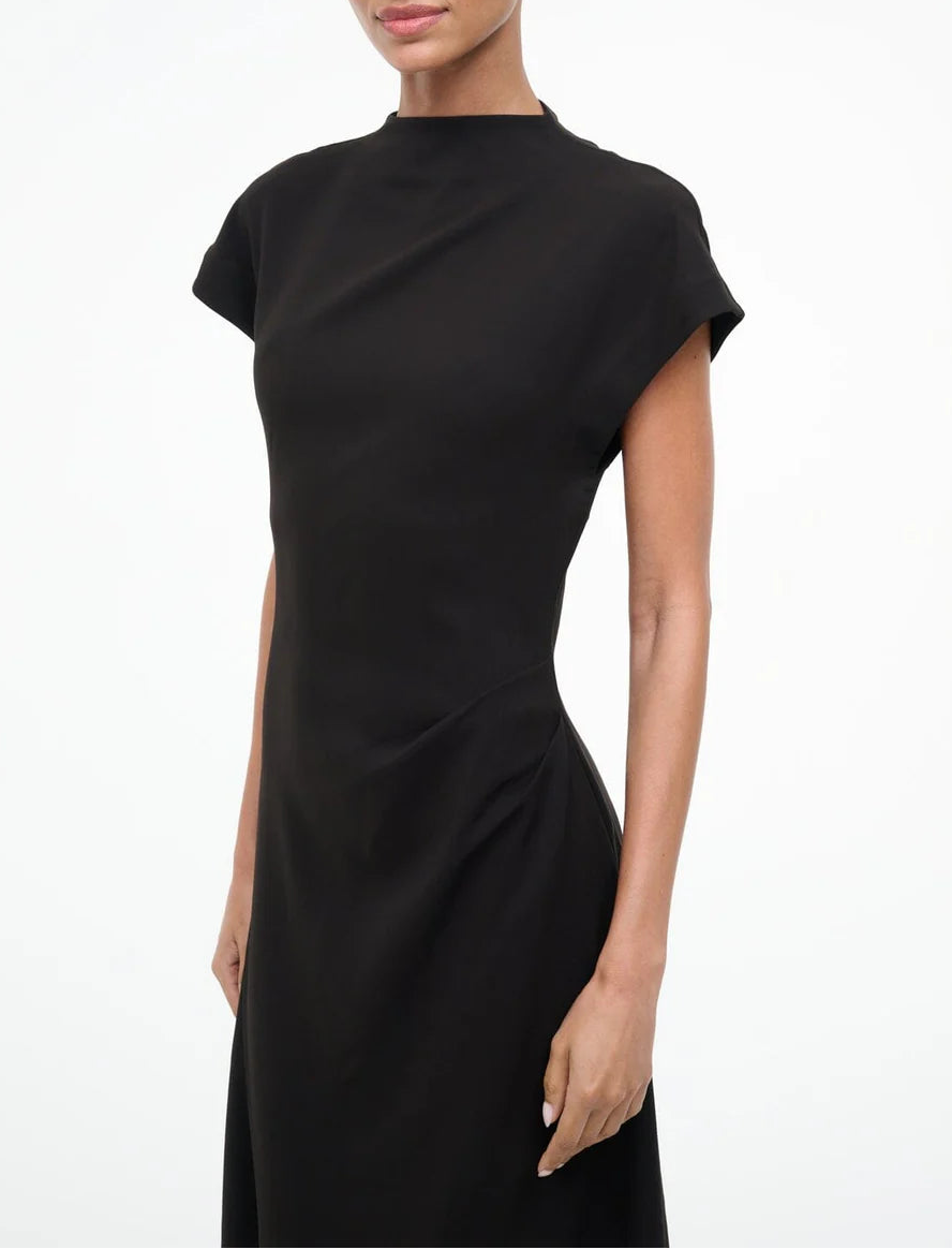 A woman stands against a plain white background wearing the Staud Zaya Dress, a black, high-neck, cap-sleeve A-line maxi made from triacetate crepe.