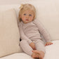 A young child with blond hair and a bow sits on a cream-colored sofa, wearing the Quincy Mae Baby Girls’ and Girls’ Waffle Top + Pant Set in light beige for a comfortable fit, looking slightly off to the side.