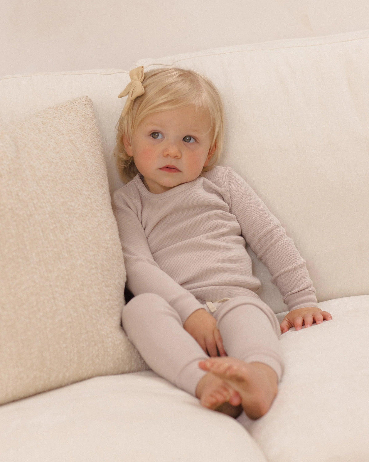 A young child with blond hair and a bow sits on a cream-colored sofa, wearing the Quincy Mae Baby Girls’ and Girls’ Waffle Top + Pant Set in light beige for a comfortable fit, looking slightly off to the side.