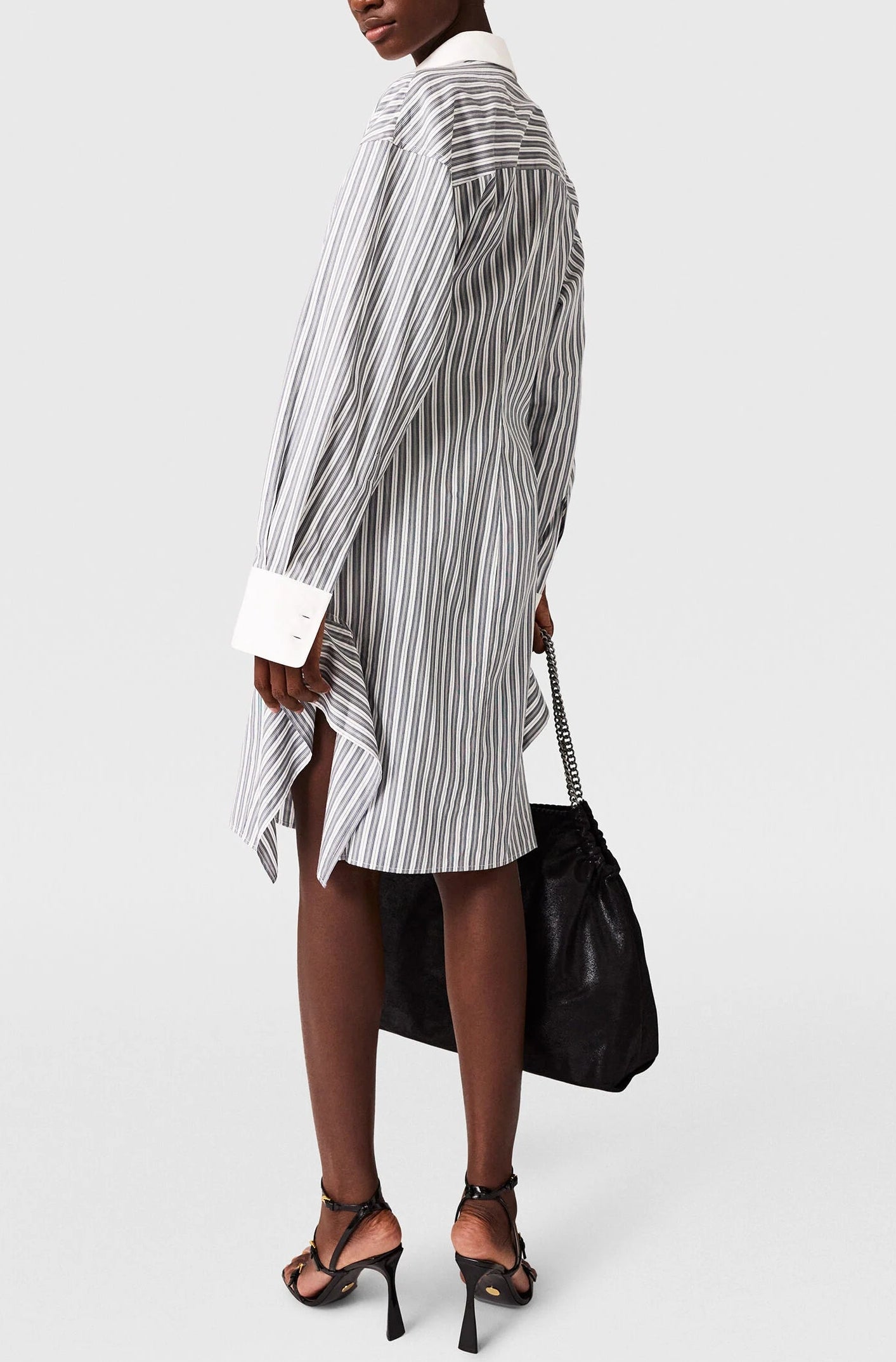 A person is shown from the back against a plain background wearing the Stella McCartney Long Sleeve Pinstripe Shirt Dress with black high-heeled sandals and holding a large black bag.