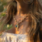 A woman in a white off-shoulder top wears Anabel Aram’s Milos Stone Drop Charm, Turquoise necklace and gold earrings, photographed in warm sunlight.