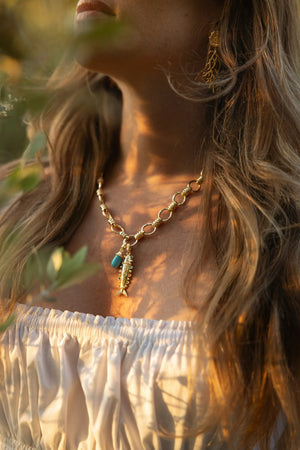 A woman in a white off-shoulder top wears Anabel Aram’s Milos Stone Drop Charm, Turquoise necklace and gold earrings, photographed in warm sunlight.