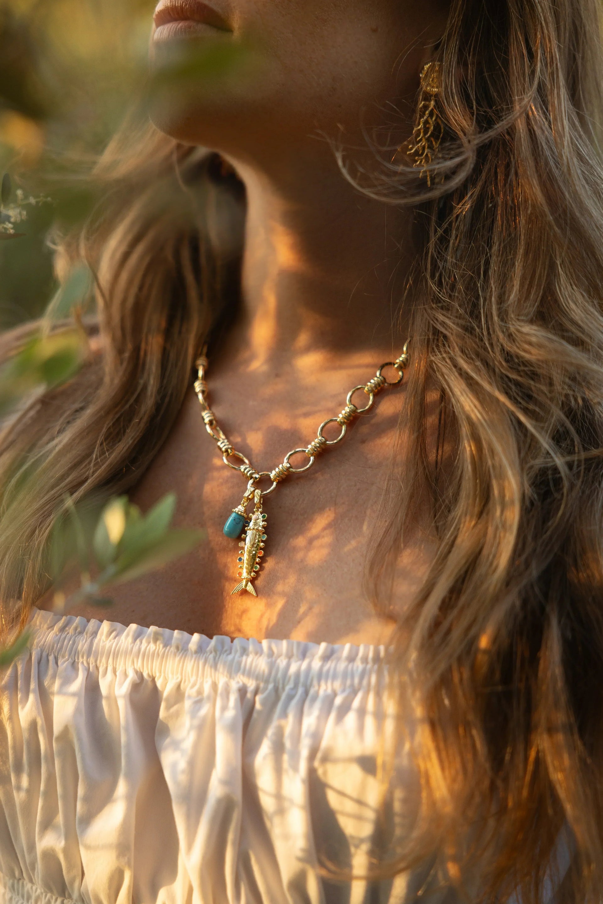 A woman in soft natural light wears Anabel Aram’s Milos Medium Fish Charm gold necklace with blue and gold pendants, matching gold earrings, and an off-the-shoulder white top.