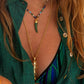 A person wears a green shirt and colorful bikini top, accessorized with layered gold necklaces—a Milos Stone Horn Charm by Anabel Aram and a beaded fish pendant—for a personalized look.