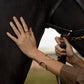 Wearing the Anabel Aram Cheval Bit Cuff, a person touches a black horse’s neck and holds its reins outdoors.