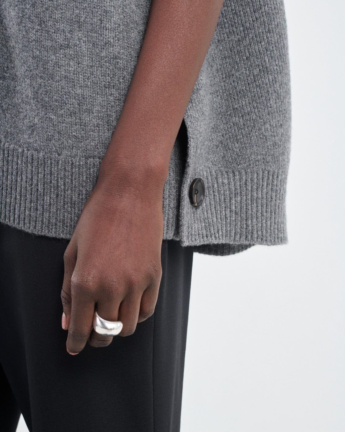 A person wearing a gray Maria McManus Cocoon Cape crafted from an alpaca-wool blend with side button detail, paired with black pants and a silver ring.
