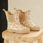 A pair of Quincy Mae Girls' Rain Boots in beige with a pink and brown floral pattern and comfy stretch panels, displayed on a light wooden stool.