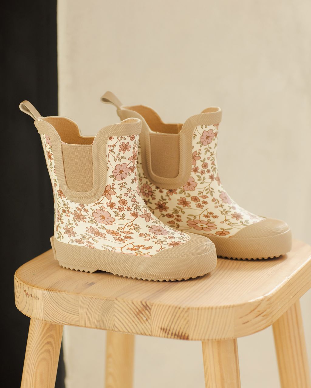 A pair of Quincy Mae Girls' Rain Boots in beige with a pink and brown floral pattern and comfy stretch panels, displayed on a light wooden stool.