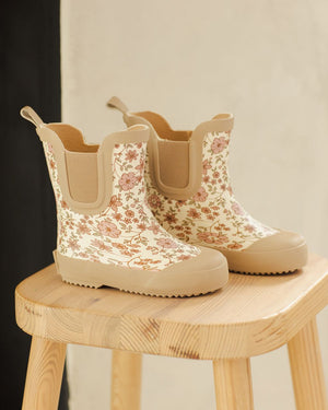 A pair of Quincy Mae Girls' Rain Boots in beige with a pink and brown floral pattern and comfy stretch panels, displayed on a light wooden stool.