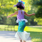 A child twirls on a sunlit park path, wearing the Misty Mermaid Dress by Great Pretenders, shimmering with iridescent sequins and paired with a purple wig.