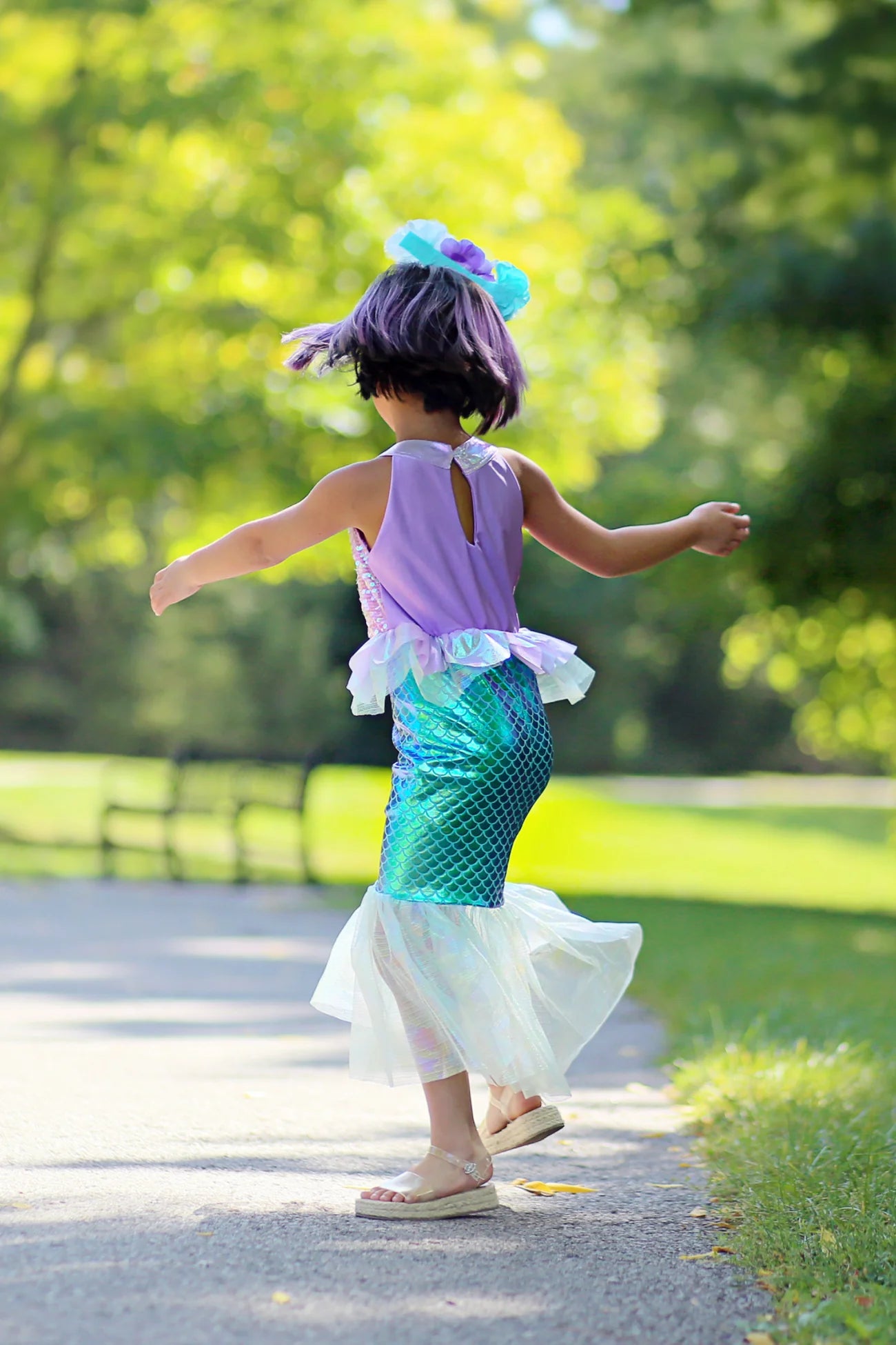 A child twirls on a sunlit park path, wearing the Misty Mermaid Dress by Great Pretenders, shimmering with iridescent sequins and paired with a purple wig.