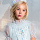 A young girl with blonde hair wears a light blue, sheer princess dress and the sparkling Snow Queen Tiara by Tutu du Monde, standing in front of a snowy, blurred background with a house visible.