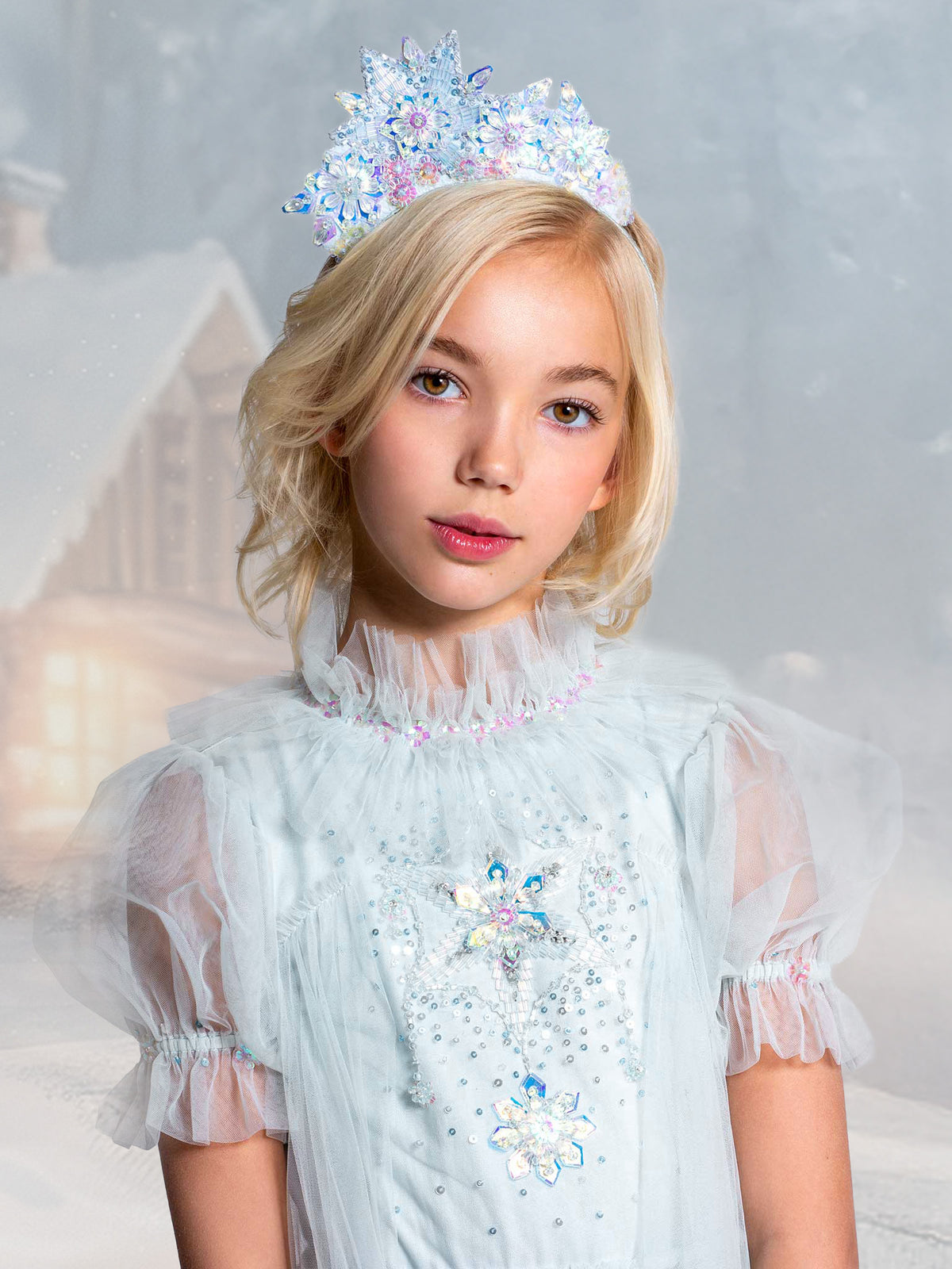 A young girl with blonde hair wears a light blue, sheer princess dress and the sparkling Snow Queen Tiara by Tutu du Monde, standing in front of a snowy, blurred background with a house visible.