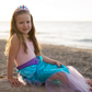 A girl in a shimmering Great Pretenders Mermaid Glimmer Skirt and Tiara sitting on a log on the beach.