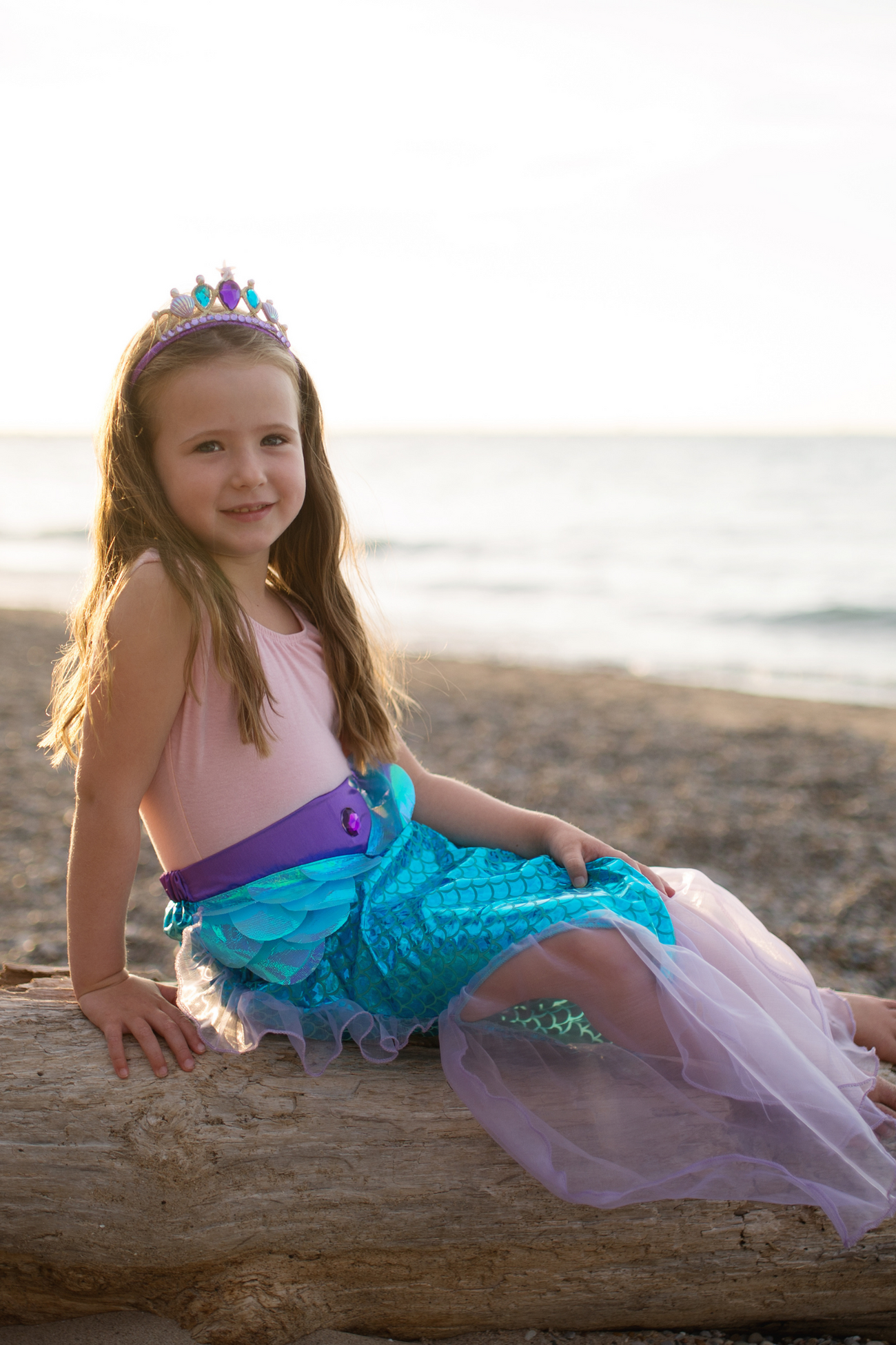 A girl in a shimmering Great Pretenders Mermaid Glimmer Skirt and Tiara sitting on a log on the beach.