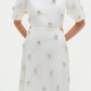 A woman wears the Simkhai Starla Embellished Midi Dress—cotton poplin with oversized short sleeves and pearl-like floral accents—standing with her hands behind her back.