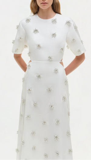 A woman wears the Simkhai Starla Embellished Midi Dress—cotton poplin with oversized short sleeves and pearl-like floral accents—standing with her hands behind her back.