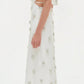 Woman in profile wears the Simkhai Starla Embellished Midi Dress, featuring a cinched waist, side cut-out, short sleeves, floral appliqués, and white heeled sandals against a plain background.