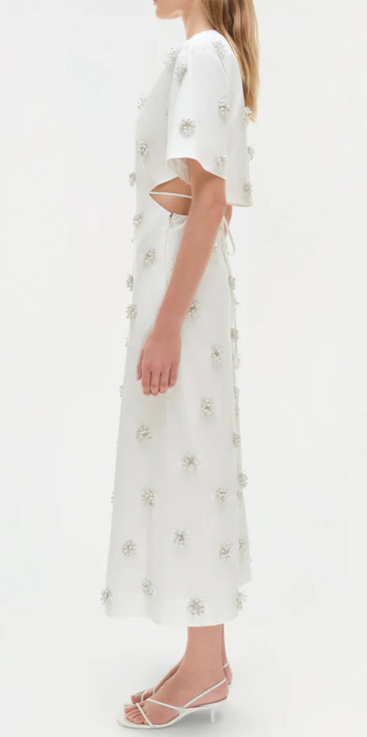 Woman in profile wears the Simkhai Starla Embellished Midi Dress, featuring a cinched waist, side cut-out, short sleeves, floral appliqués, and white heeled sandals against a plain background.