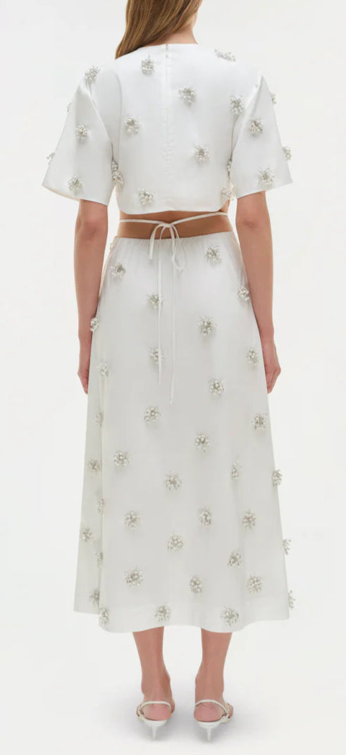 A woman faces away in the Simkhai Starla Embellished Midi Dress—white cotton poplin with textured floral details and a cinched waist—paired with white sandals.