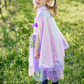 A young girl in a Great Pretenders Sequins Cape and a purple dress, holding a bouquet, stands in a blooming orchard, looking over her shoulder with a smile.