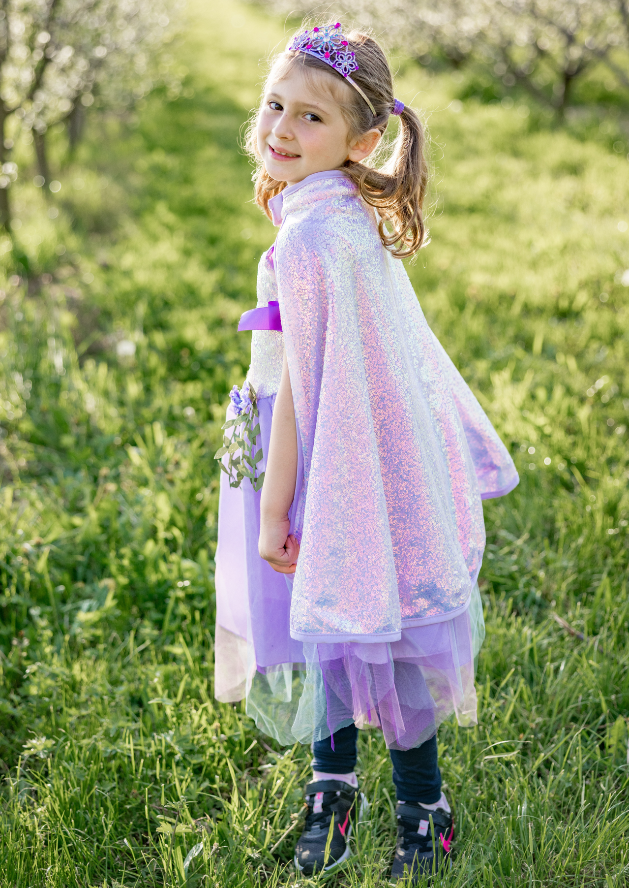 A young girl in a Great Pretenders Sequins Cape and a purple dress, holding a bouquet, stands in a blooming orchard, looking over her shoulder with a smile.