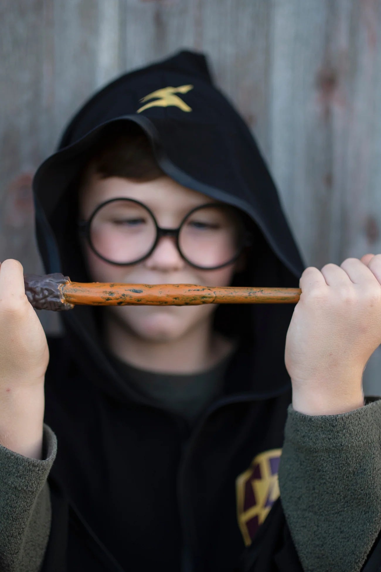 A child dressed in a black hooded robe and round glasses holds the Great Pretenders Wizard Wand horizontally in front of their face, making it the perfect kids costume accessory.