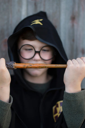 A child dressed in a black hooded robe and round glasses holds the Great Pretenders Wizard Wand horizontally in front of their face, making it the perfect kids costume accessory.