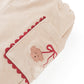 A close-up of the Baby Girls’ Nellie Pant with Poodle Pocket by Gingersnaps shows beige fabric pants with an elastic waistband, playful poodle-embroidered pocket accented with red trim, and comfy straight-leg styling for charming everyday wear.