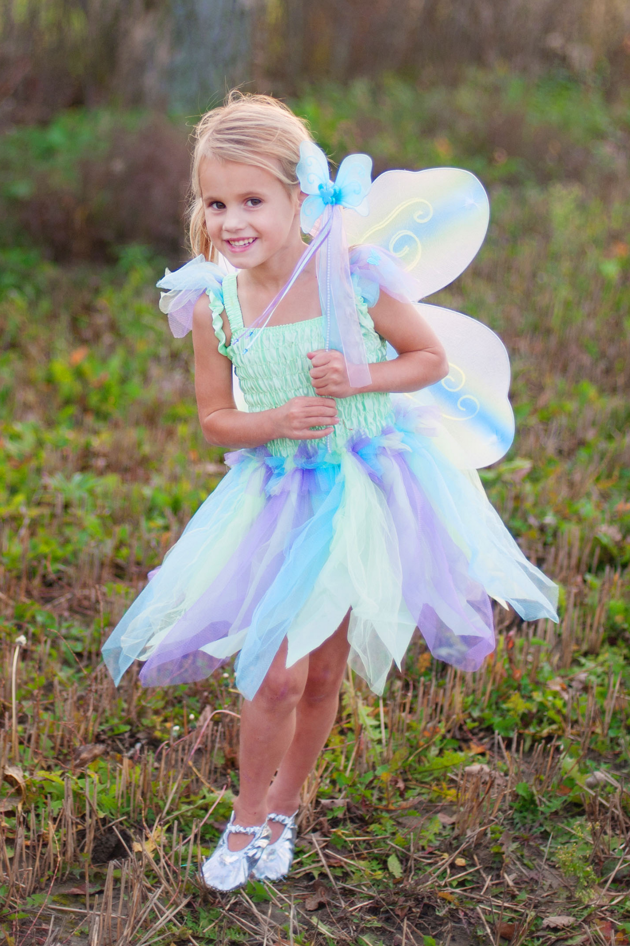 A young girl dressed in a Great Pretenders Butterfly Dress with Wings and Wand, smiling in an outdoor setting.