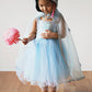 A young child wearing the Great Pretenders Snow Queen Cape, adorned with silver glitter stars, stands barefoot on a wooden floor holding a pink flower wand and looking down.