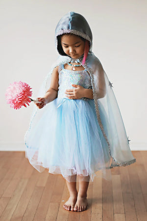 A young child wearing the Great Pretenders Snow Queen Cape, adorned with silver glitter stars, stands barefoot on a wooden floor holding a pink flower wand and looking down.