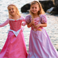 Two young girls dressed in Great Pretenders Boutique Princess Gowns are standing by a lakeside, smiling and holding hands.