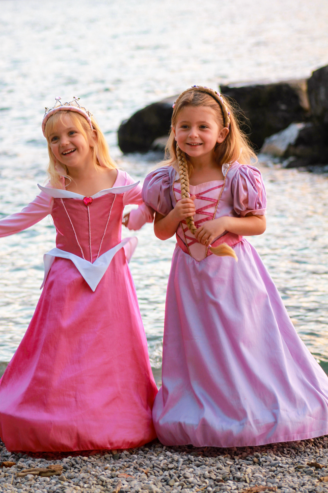 Two young girls dressed in Great Pretenders Boutique Princess Gowns are standing by a lakeside, smiling and holding hands.