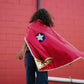A child wearing the Great Pretenders Gold & Red Reversible Wonder Cape stands in front of a red brick wall, facing away from the camera.