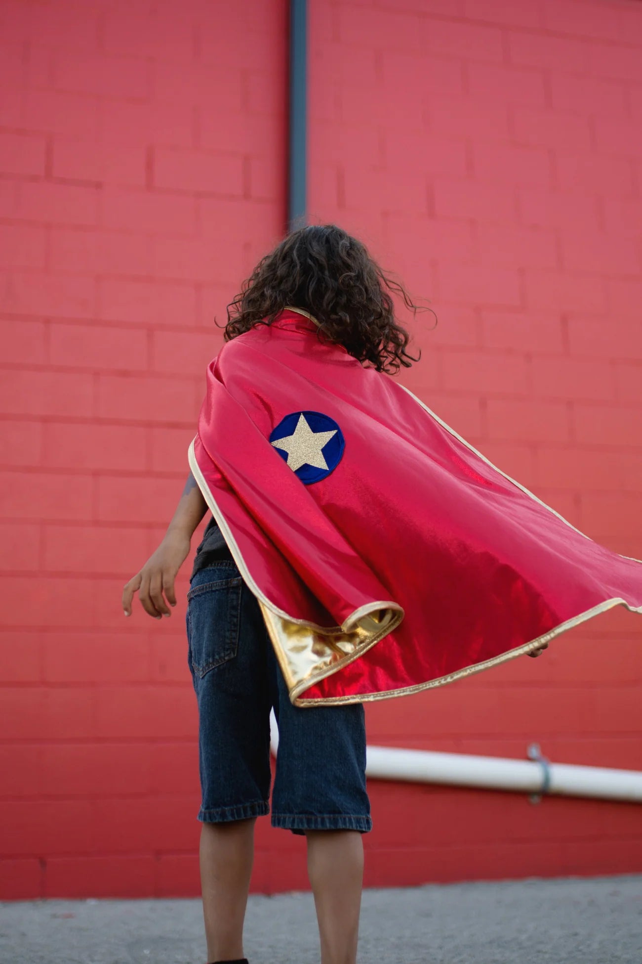 A child wearing the Great Pretenders Gold & Red Reversible Wonder Cape stands in front of a red brick wall, facing away from the camera.
