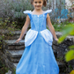 A young girl stands on stone steps outdoors, smiling while dressed in a blue princess costume from the Great Pretenders Boutique Princess Gown Collection with a tiara.